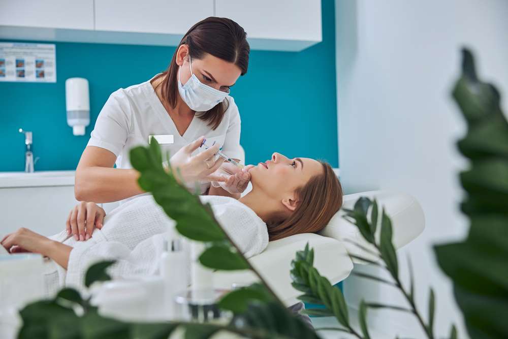 Brunette beautician in uniform holding syringe with an injection needle for nourishing and rejuvenating the skin while patient lying on medicine chair in beauty salon